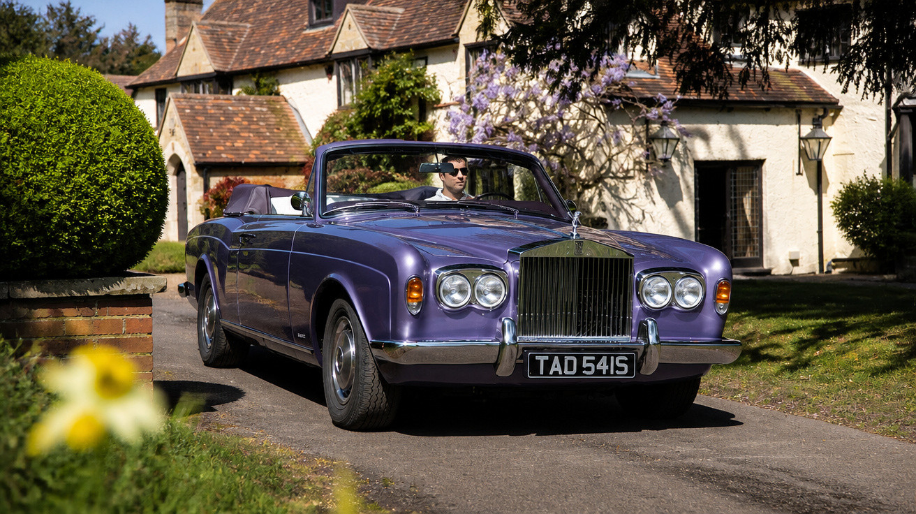 Carro conversível clássico roxo parado em frente a casa antiga com jardim ao redor.