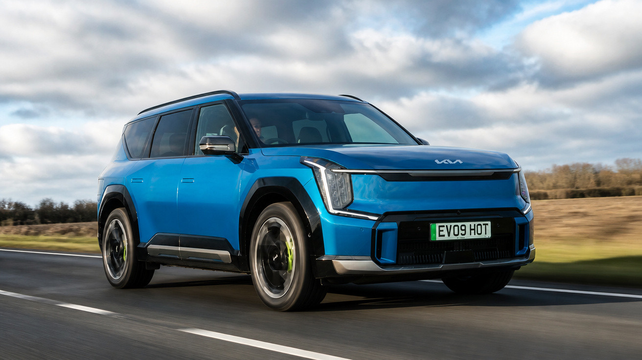 Carro SUV elétrico azul em movimento em estrada asfaltada sob céu nublado e campo ao fundo.