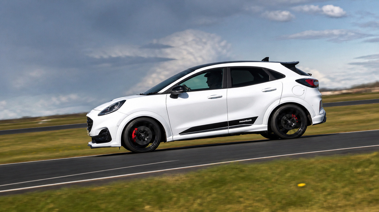 Carro branco esportivo em alta velocidade na pista com céu nublado ao fundo.