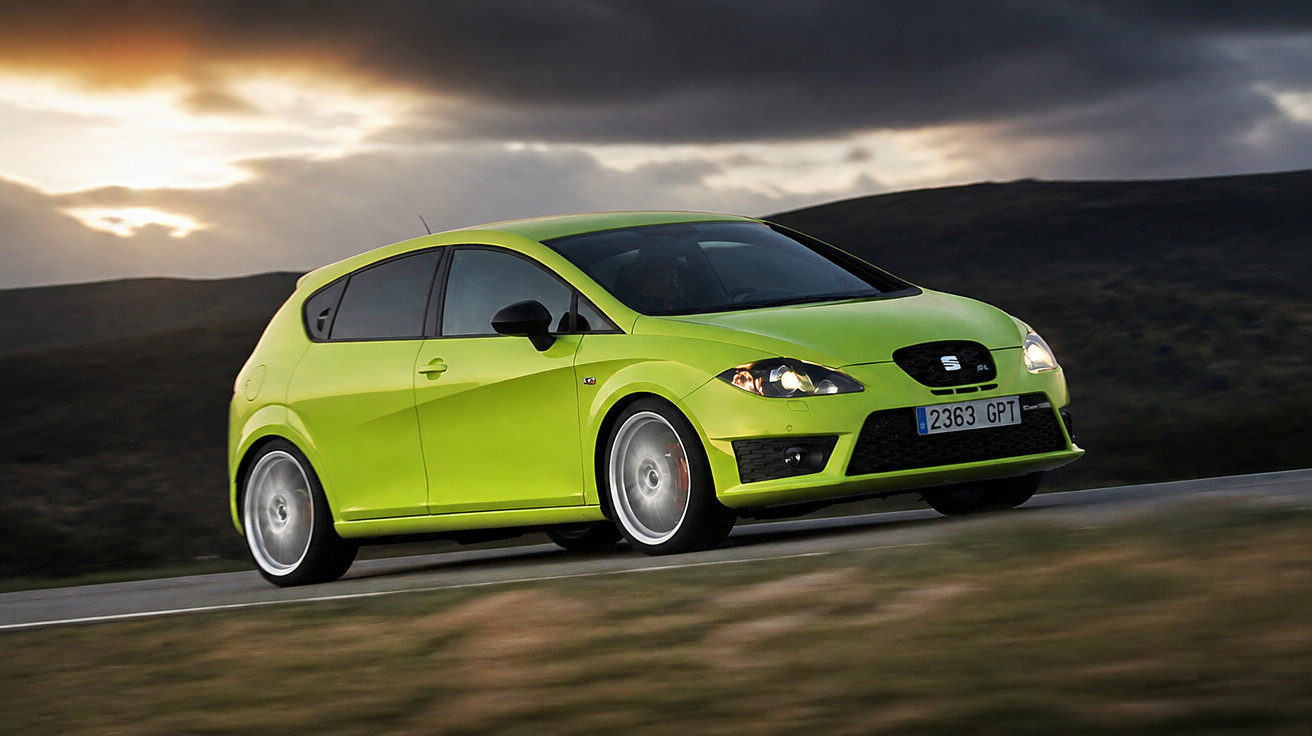 Carro hatchback verde-limão em movimento em estrada com céu nublado ao fundo.