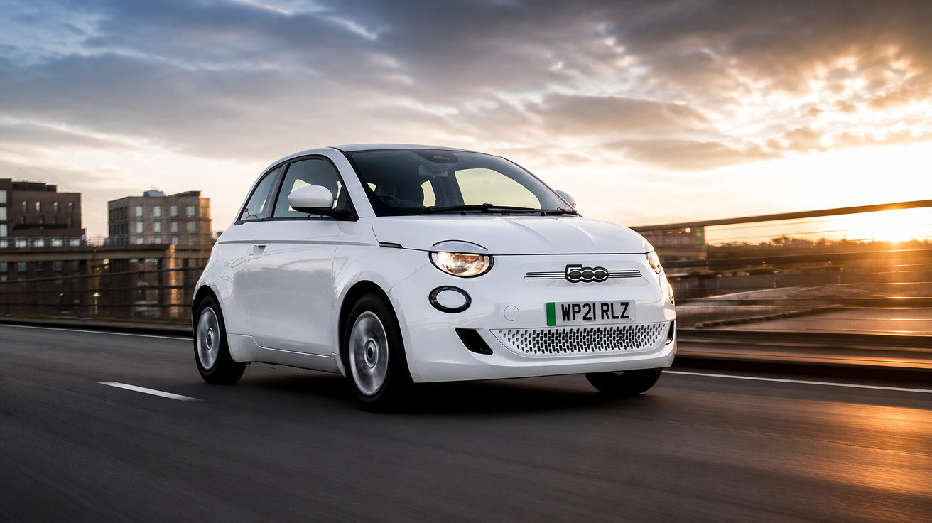 Carro branco compacto Fiat 500 em movimento na estrada ao entardecer com prédios ao fundo.