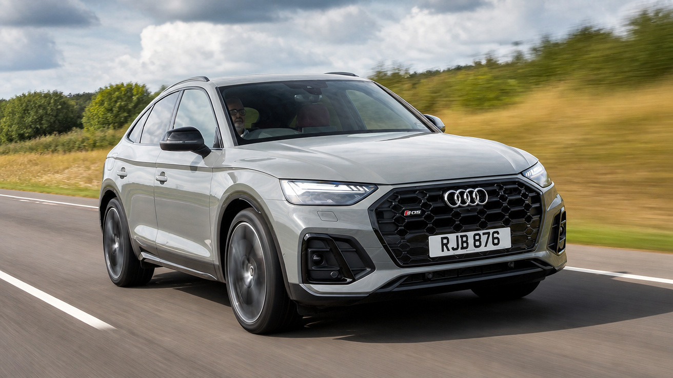 Audi Q5 cinza em movimento em estrada com vegetação ao fundo em dia nublado.