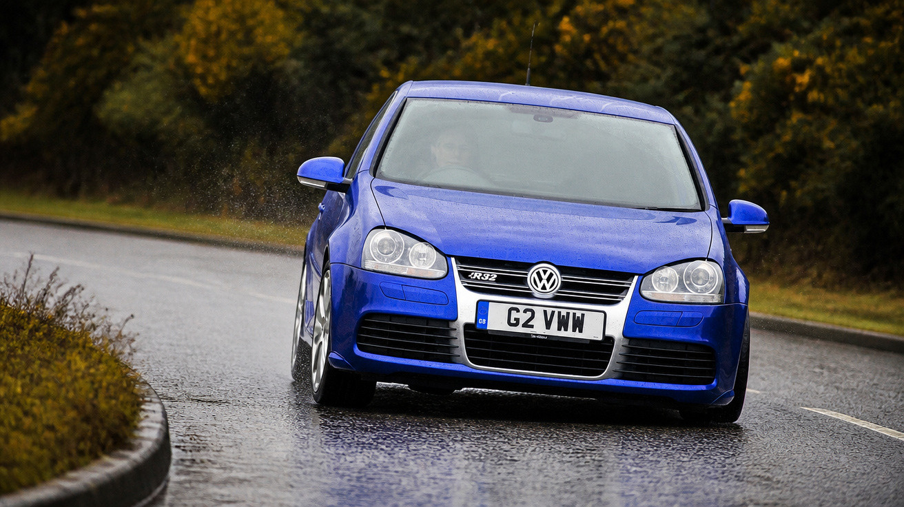 Carro esportivo azul Volkswagen Golf R32 em curva de estrada molhada cercada por vegetação.