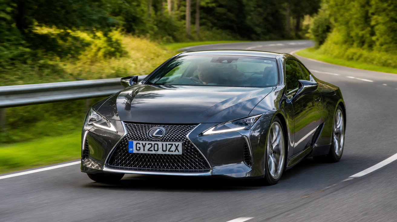 Carro esportivo cinza Lexus em estrada sinuosa cercada por árvores verdes.