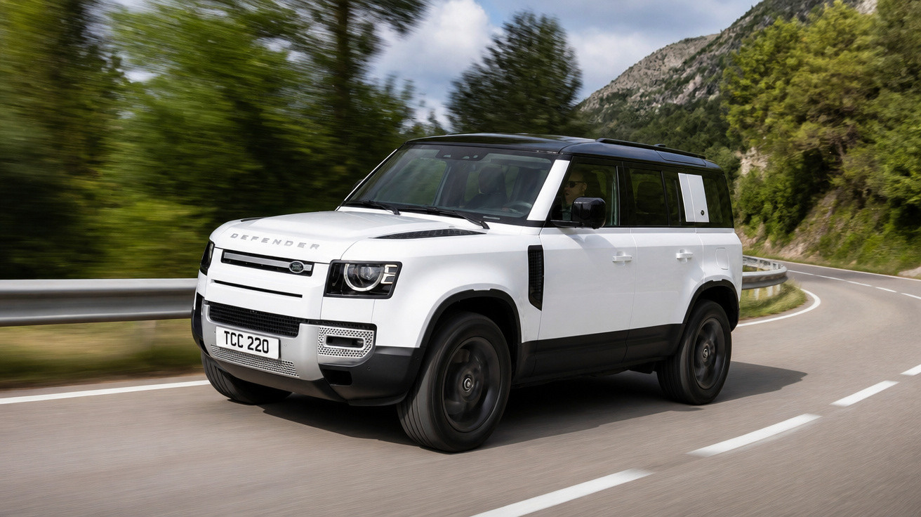 SUV branco Land Rover Defender em estrada sinuosa cercada por árvores e montanhas.