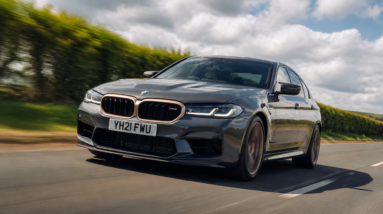Carro BMW cinza escuro em movimento em estrada com vegetação ao fundo em dia nublado.