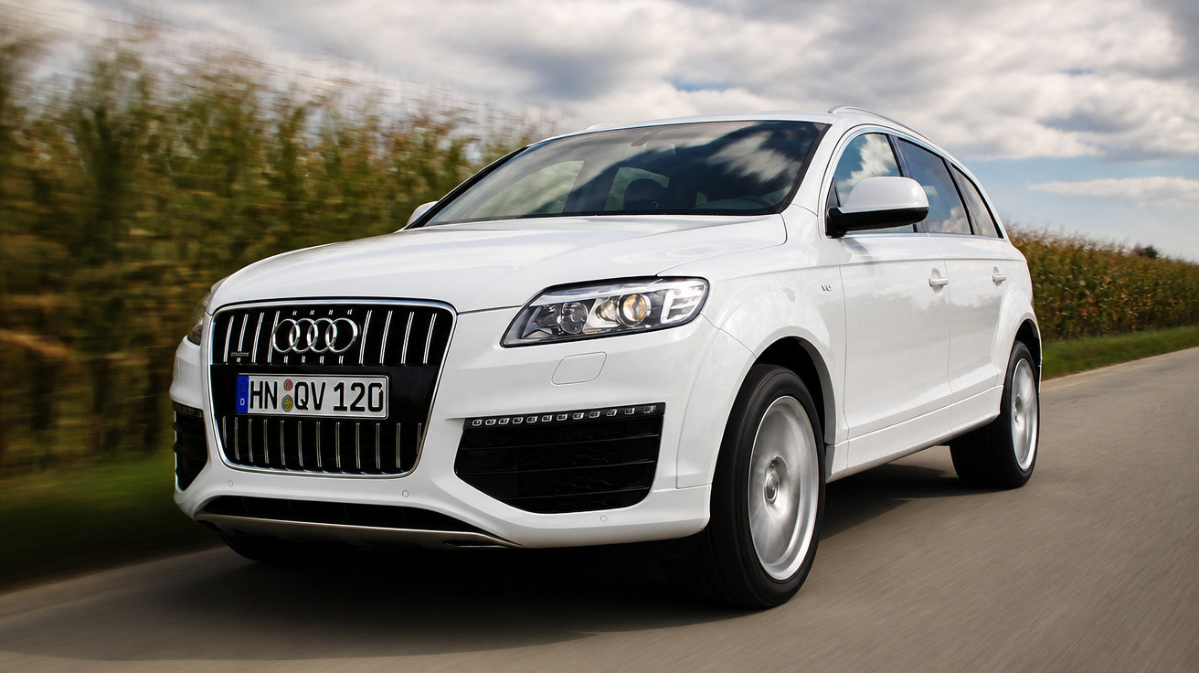 Carro branco Audi Q7 em movimento em estrada rural com céu nublado e plantações ao fundo.