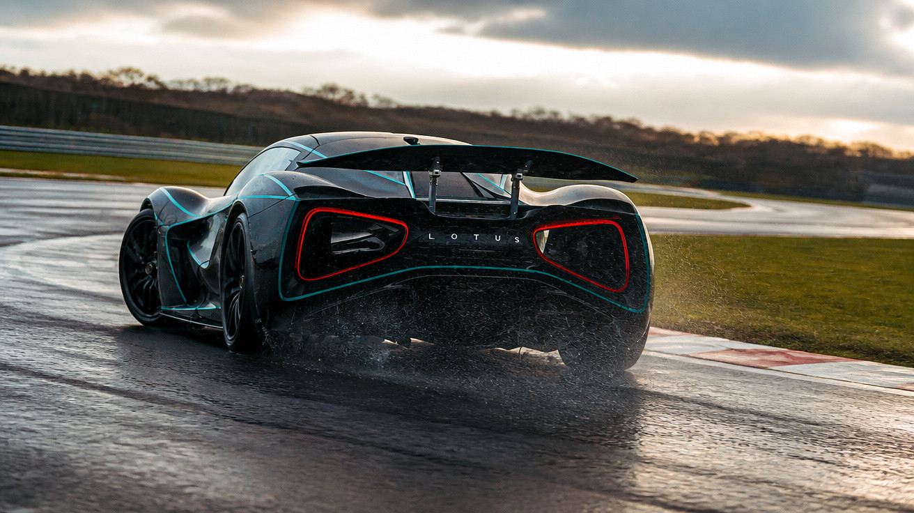 Carro esportivo preto da Lotus em pista molhada com céu nublado ao fundo.