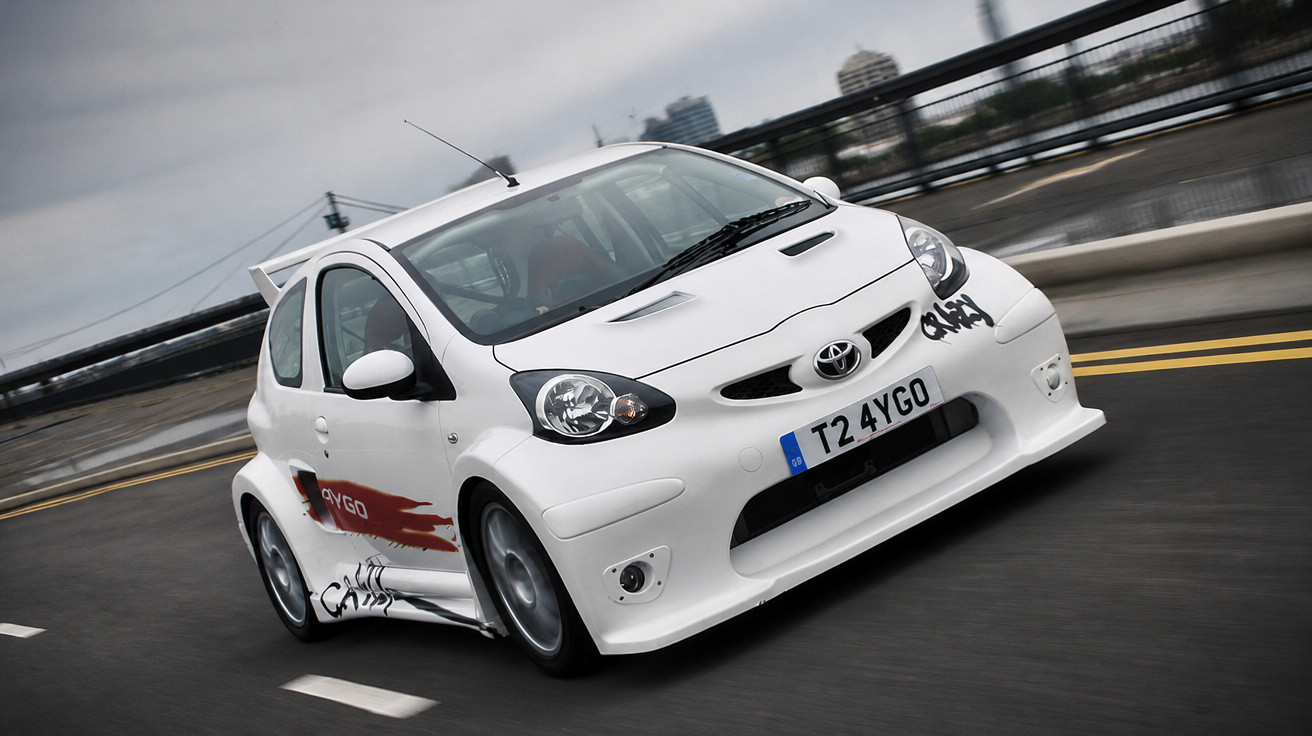 Carro branco esportivo Toyota Aygo em movimento em uma estrada urbana com faixas duplas amarelas.