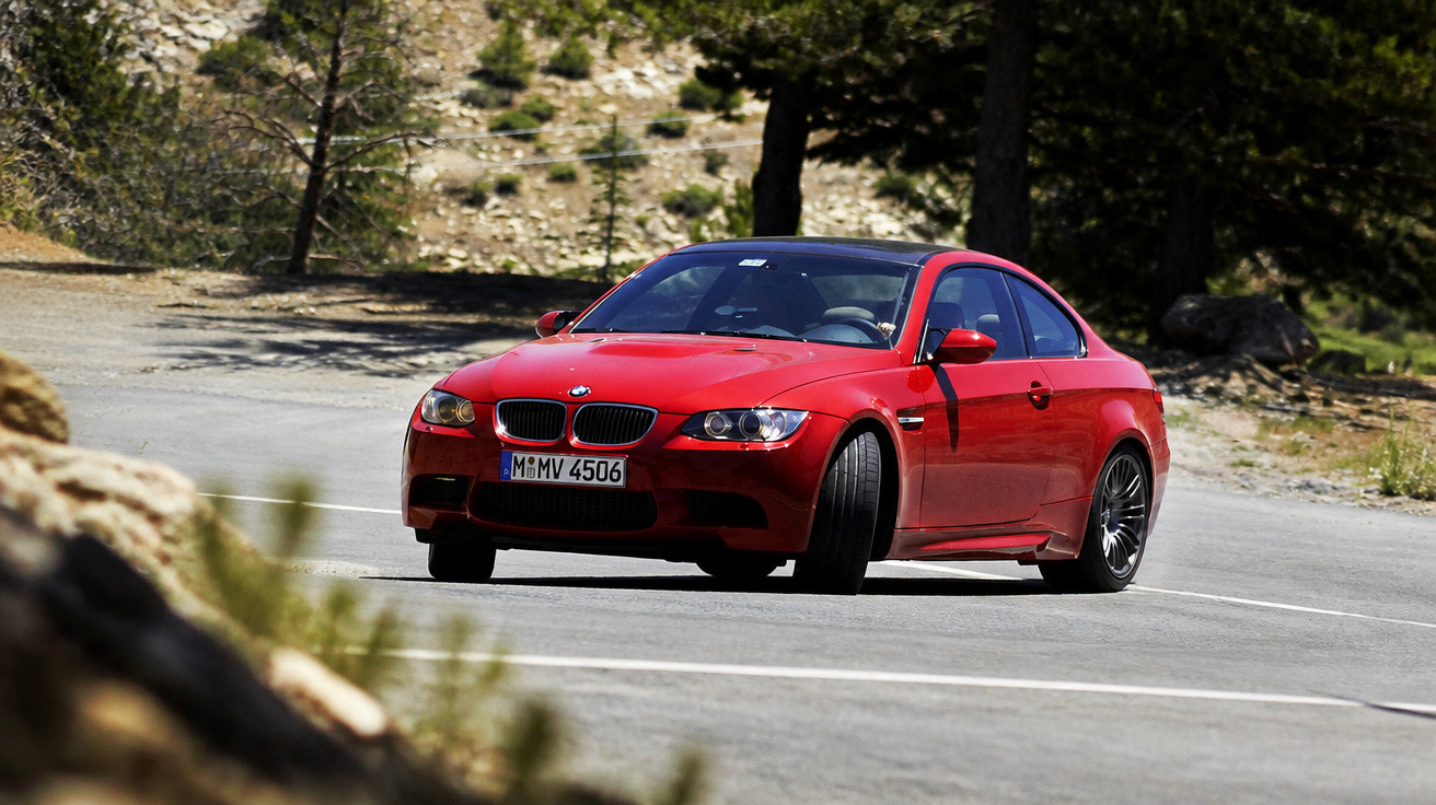 Carro esportivo BMW vermelho em curva de estrada asfaltada em área montanhosa com vegetação.