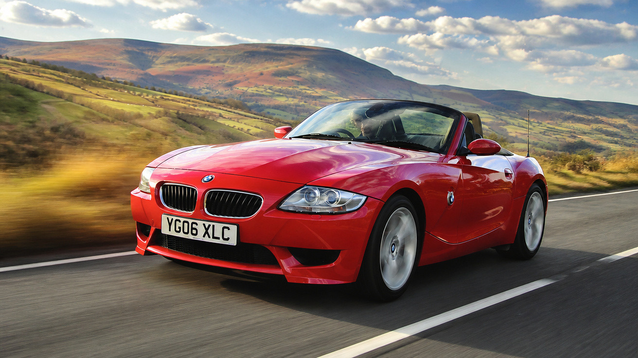 Carro esportivo conversível vermelho BMW em estrada com colinas ao fundo e céu parcialmente nublado.