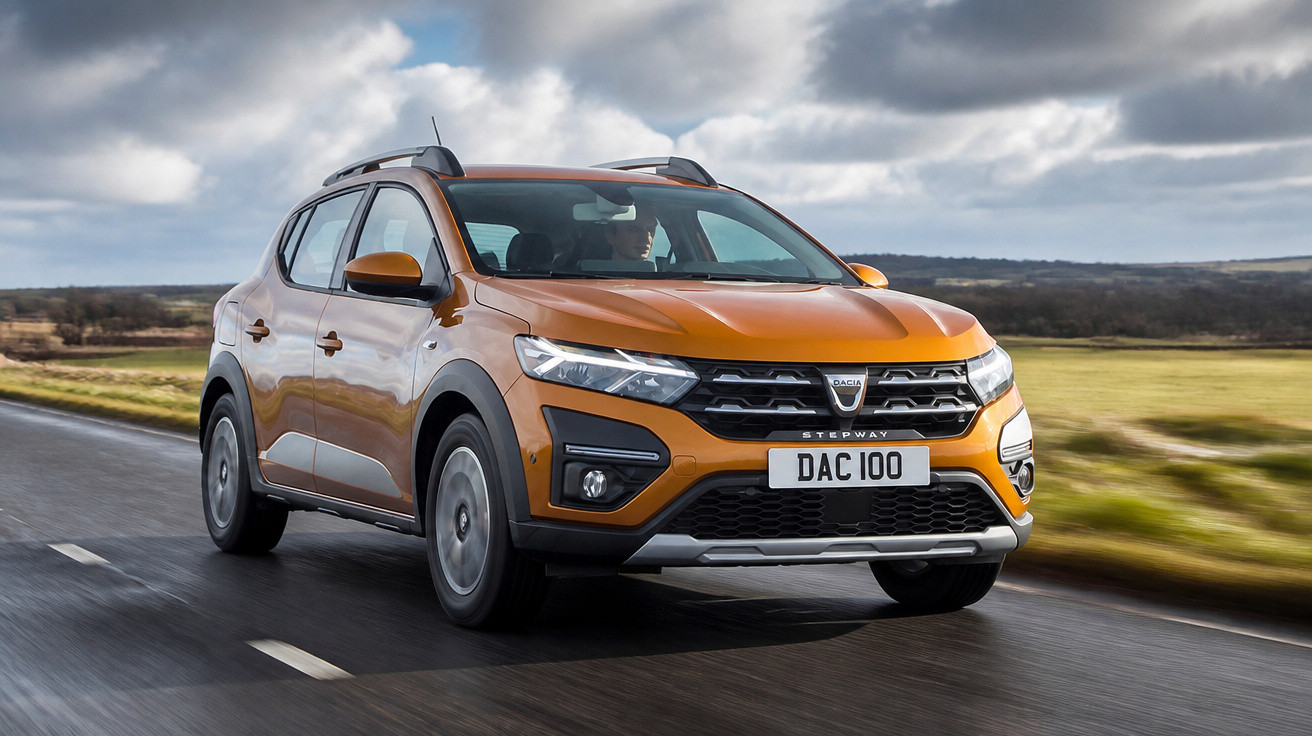 Carro SUV Dacia Stepway laranja em estrada molhada, céu nublado e campo ao fundo.