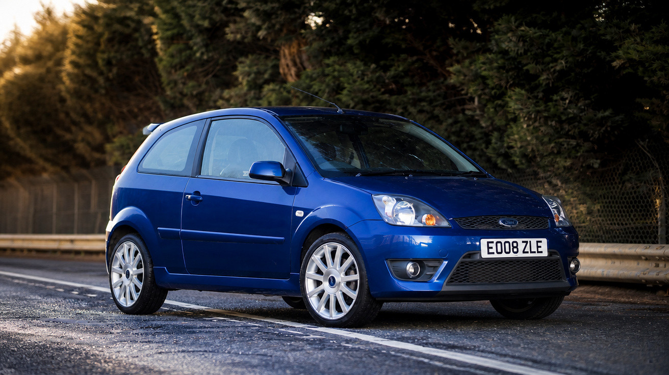 Carro hatchback azul estacionado ao lado de estrada com árvores ao fundo.