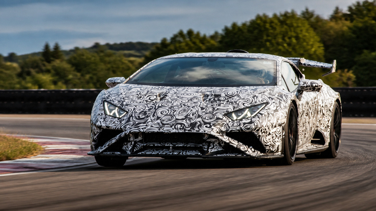Carro esportivo com camuflagem preta e branca em curva de pista de corrida cercada por árvores.
