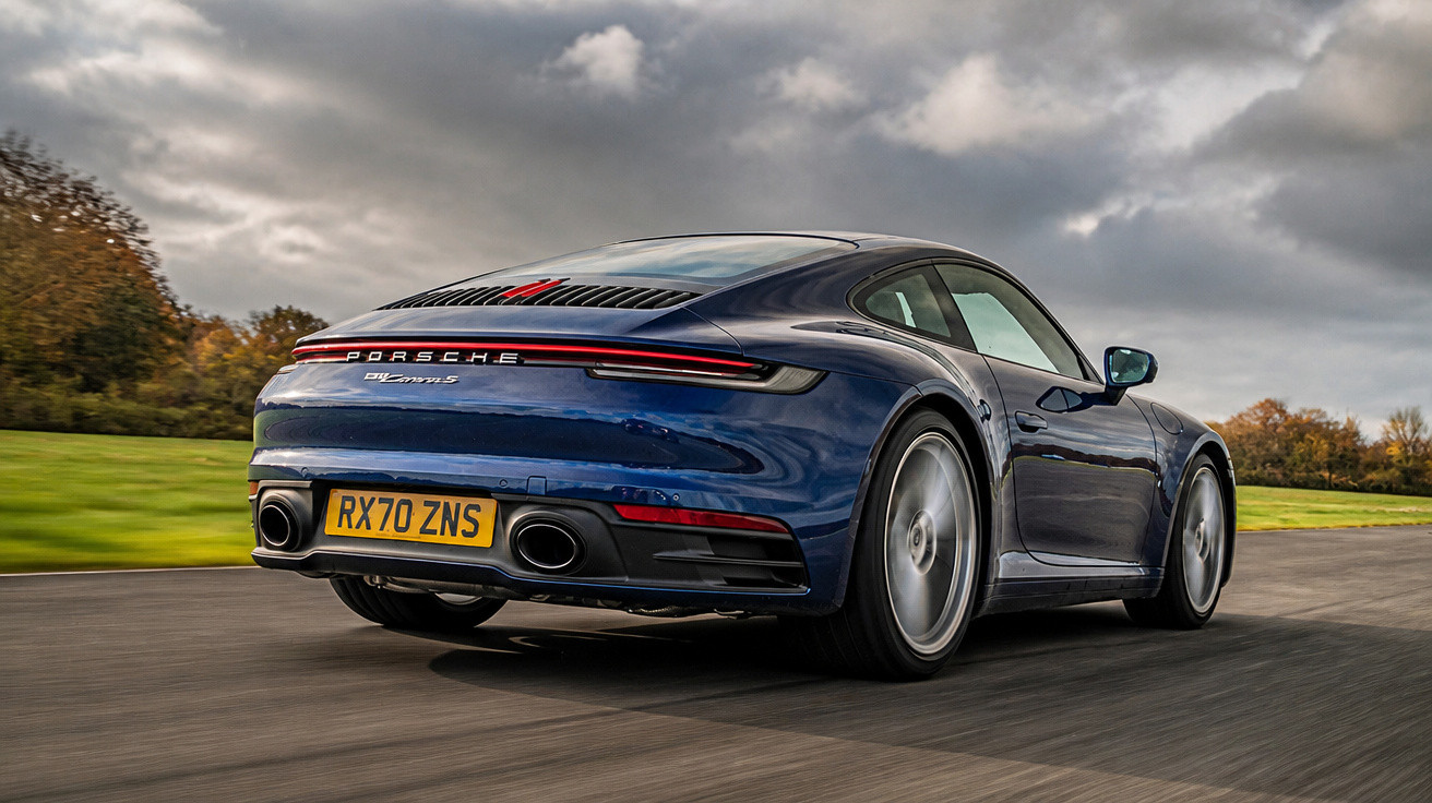 Carro esportivo Porsche 911 Carrera S azul em movimento em estrada asfaltada com céu nublado.