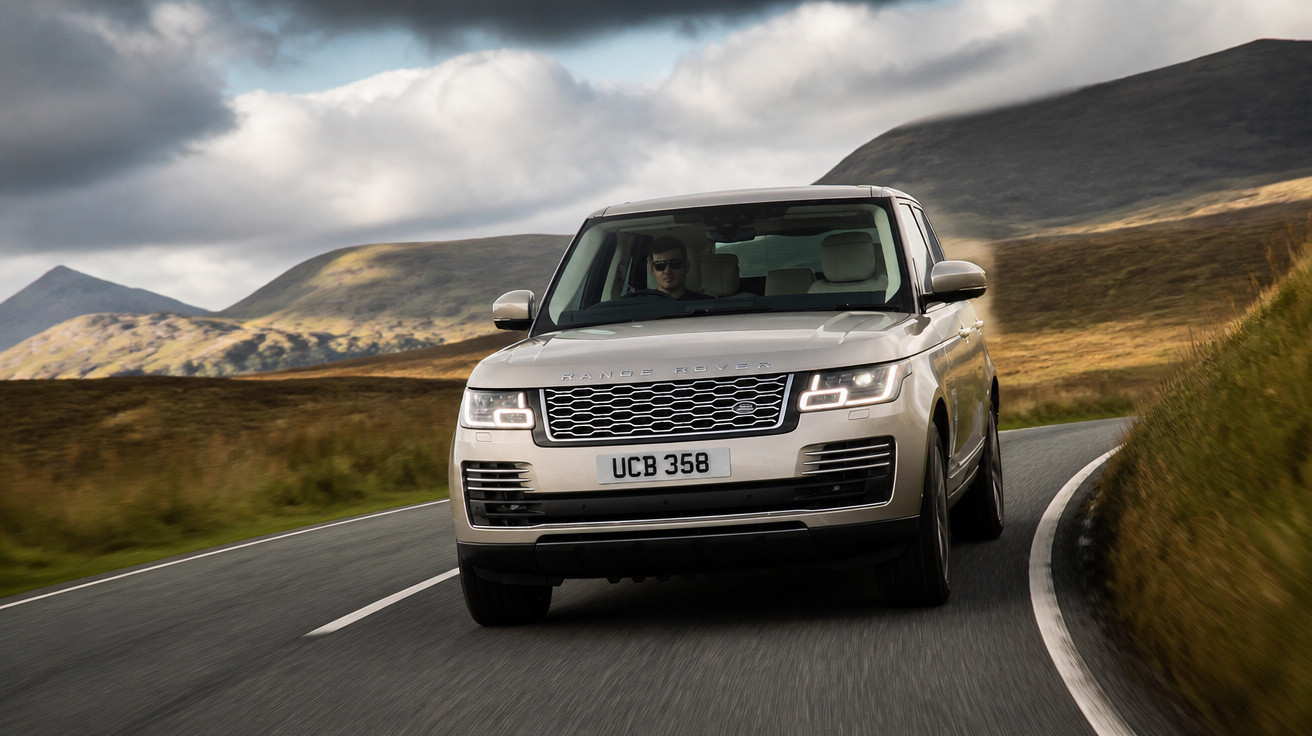 SUV prata Range Rover dirigindo em estrada sinuosa com colinas e céu nublado ao fundo.