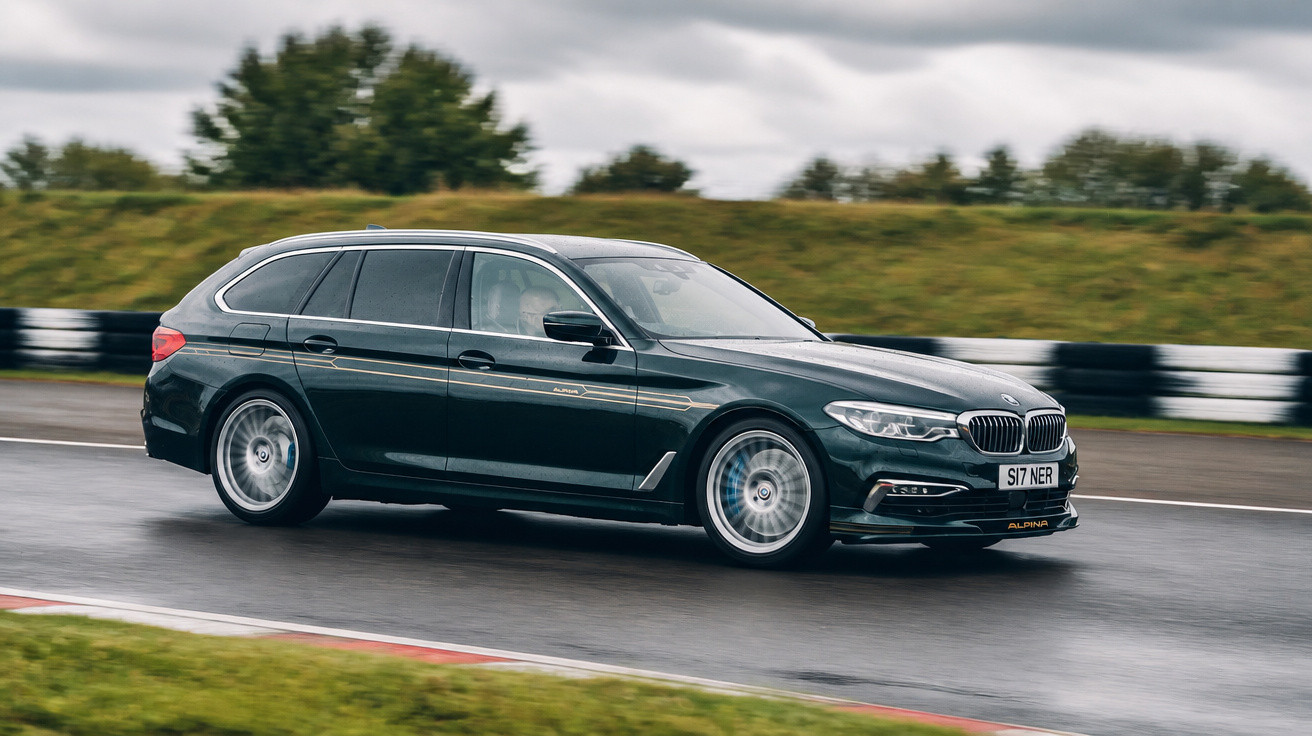 Carro BMW Alpina Touring verde escuro em pista molhada com fundo de grama e céu nublado.