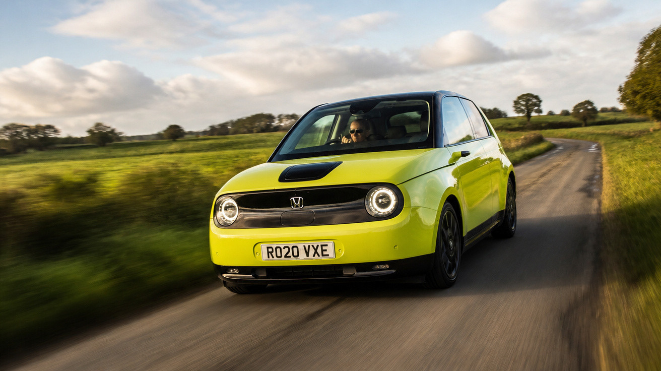 Carro compacto elétrico amarelo verde trafegando em estrada rural ao entardecer com céu claro e nuvens.