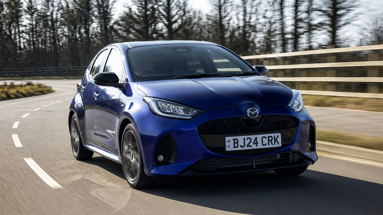 Carro hatchback azul em alta velocidade em estrada curva com árvores ao fundo em dia claro.