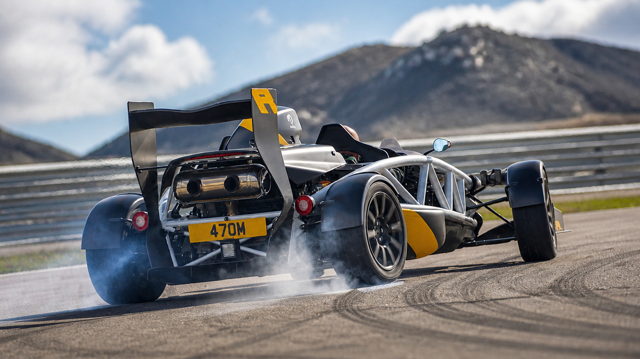 Carro esportivo preto e amarelo derrapando em pista de corrida com montanhas ao fundo.