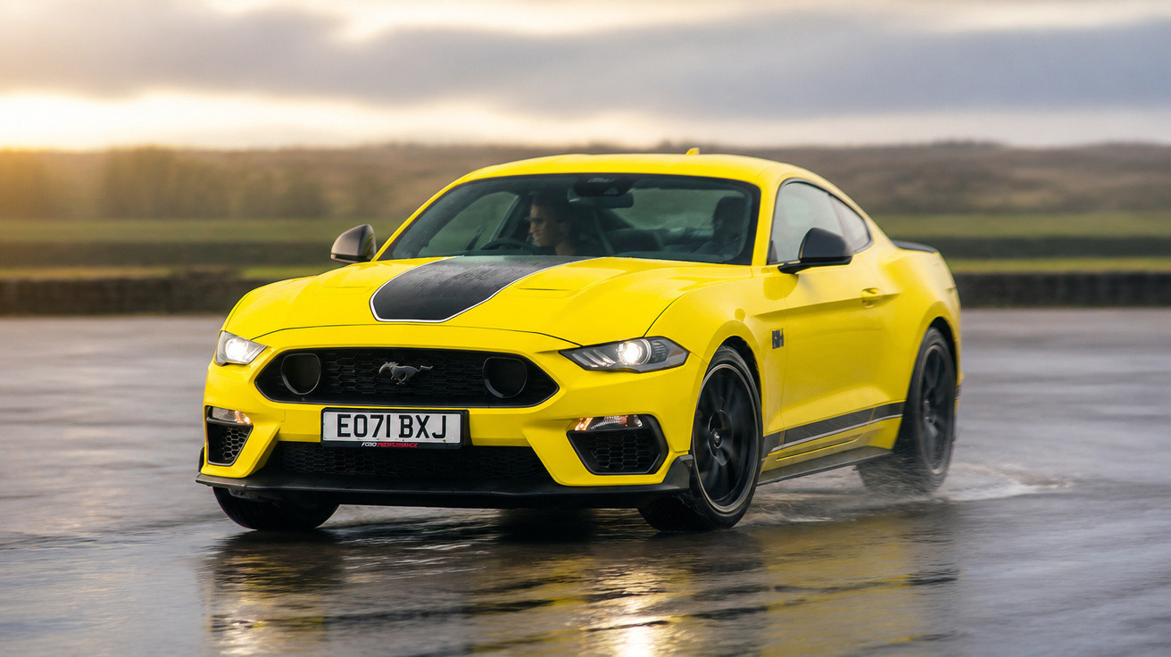 Carro esportivo amarelo Ford Mustang em pista molhada ao entardecer.