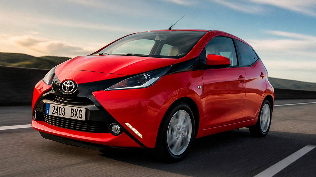Carro vermelho Toyota Aygo em movimento em uma estrada com céu azul ao fundo.