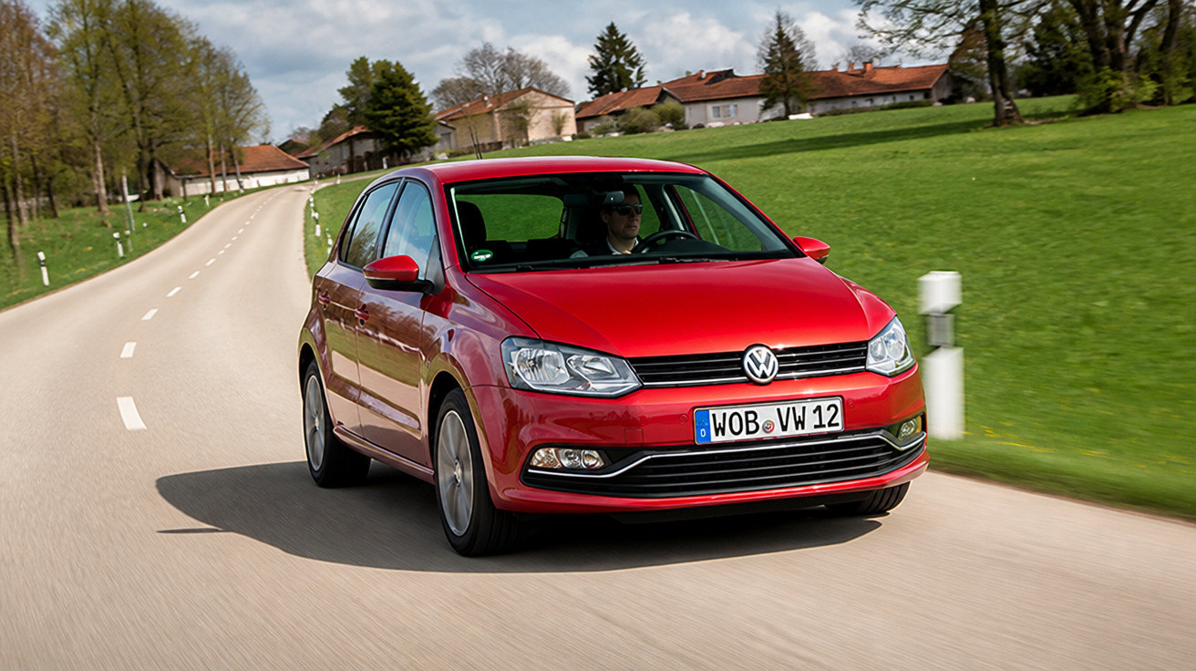 Carro vermelho Volkswagen Polo dirigido em estrada rural com casas ao fundo e gramado verde.