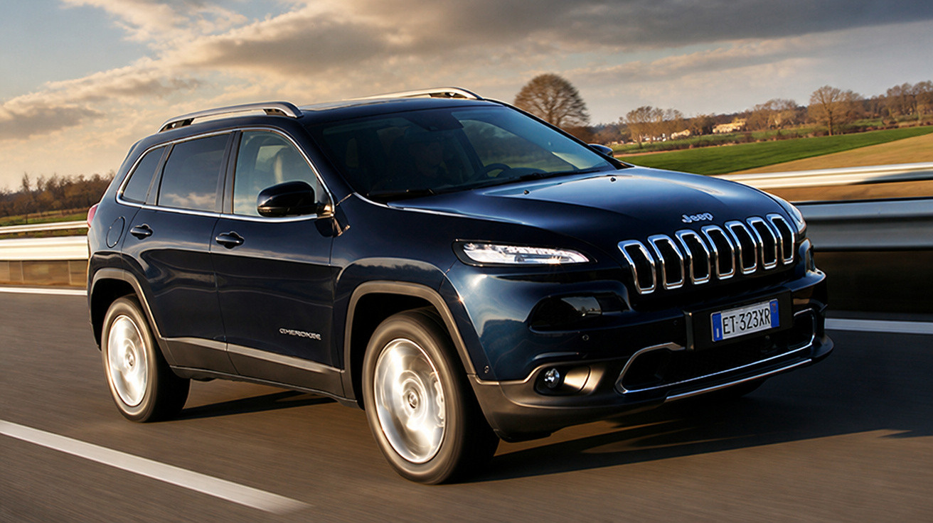 Jeep Cherokee preto em estrada asfaltada com paisagem rural ao fundo e céu parcialmente nublado.