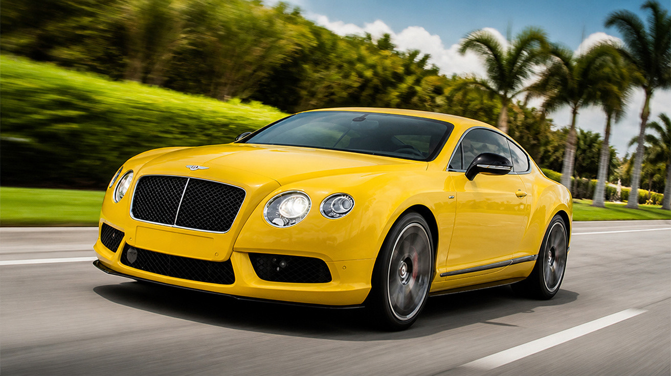 Carro esportivo amarelo Bentley em movimento em estrada com árvores e palmeiras ao fundo.