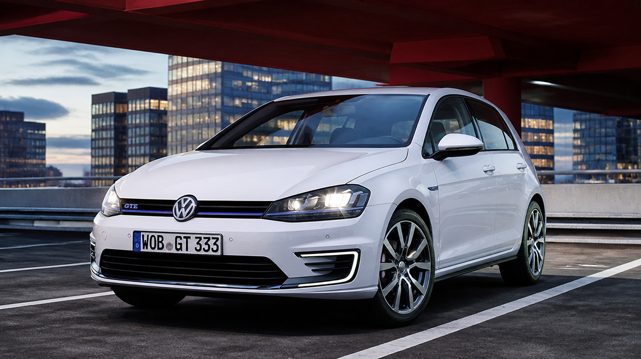 Carro branco Volkswagen Golf GTE estacionado em garagem com edifícios ao fundo ao entardecer.