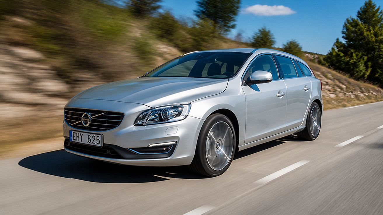 Carro prata modelo station wagon da Volvo em movimento em estrada asfaltada cercada por árvores.