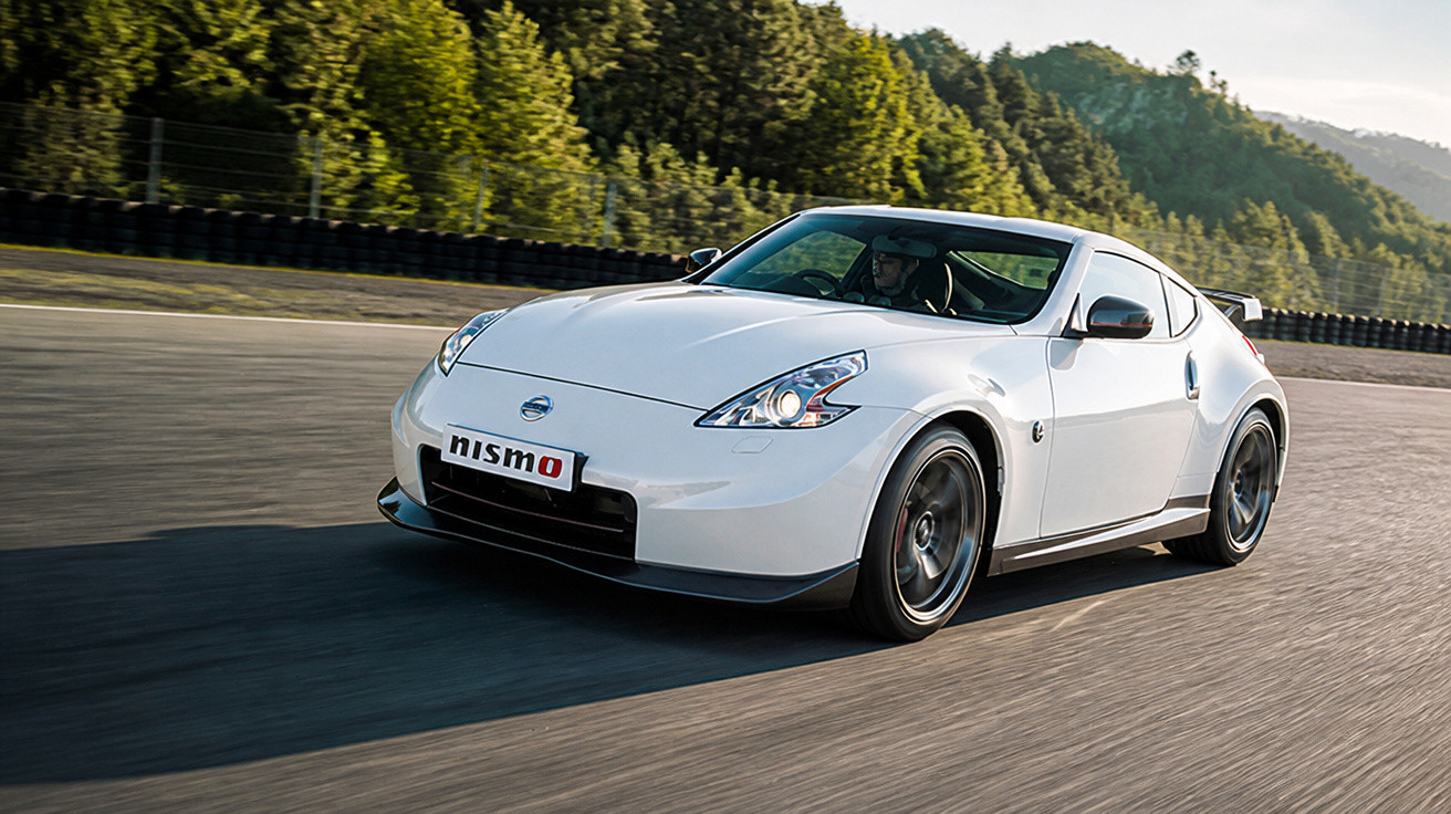 Carro esportivo branco Nissan 370Z Nismo em alta velocidade na pista com floresta ao fundo.