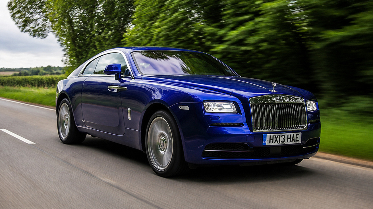 Carro esportivo azul da marca Rolls-Royce dirigindo em estrada cercada por vegetação verde.