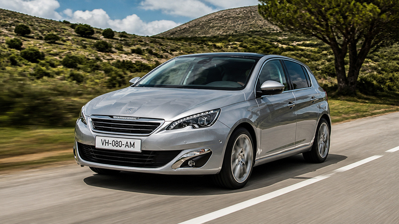 Carro prata Peugeot 308 hatch em movimento em estrada rural com colinas e árvores ao fundo.