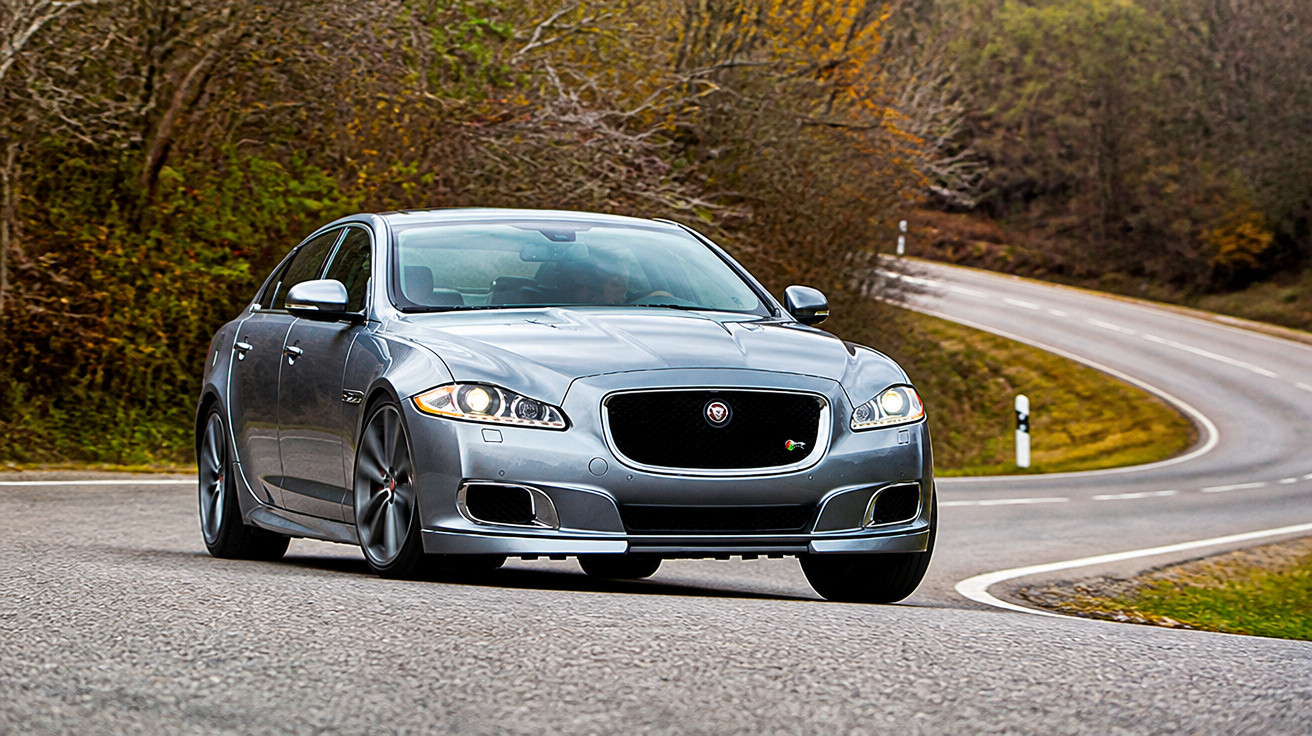 Carro cinza Jaguar sedan em curva na estrada com árvores e paisagem outonal ao fundo.