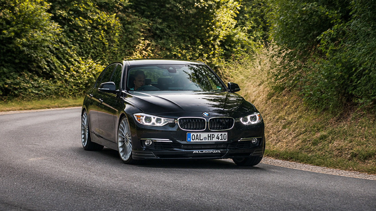 Carro BMW preto modelo Alpina em curva de estrada cercada por vegetação verde.