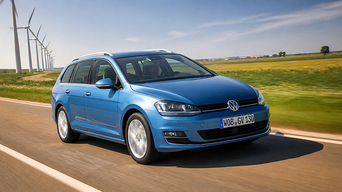 Carro Volkswagen azul em estrada com campo verde e turbinas eólicas ao fundo em dia ensolarado.