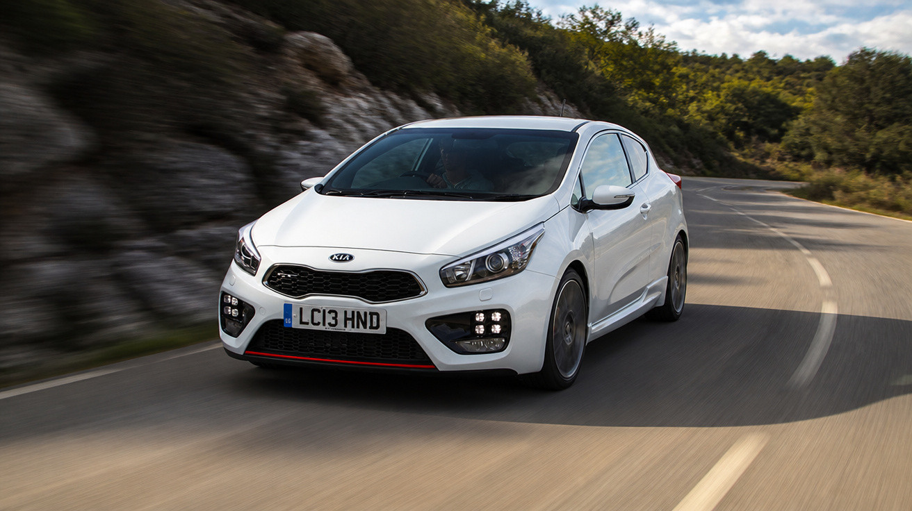 Carro branco Kia em movimento em estrada curvada rodeada por árvores e pedras.