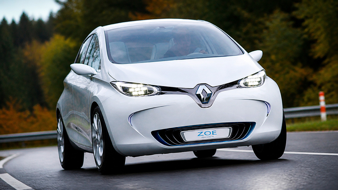 Carro elétrico branco Renault Zoe em estrada sinuosa com árvores ao fundo e faróis acesos.