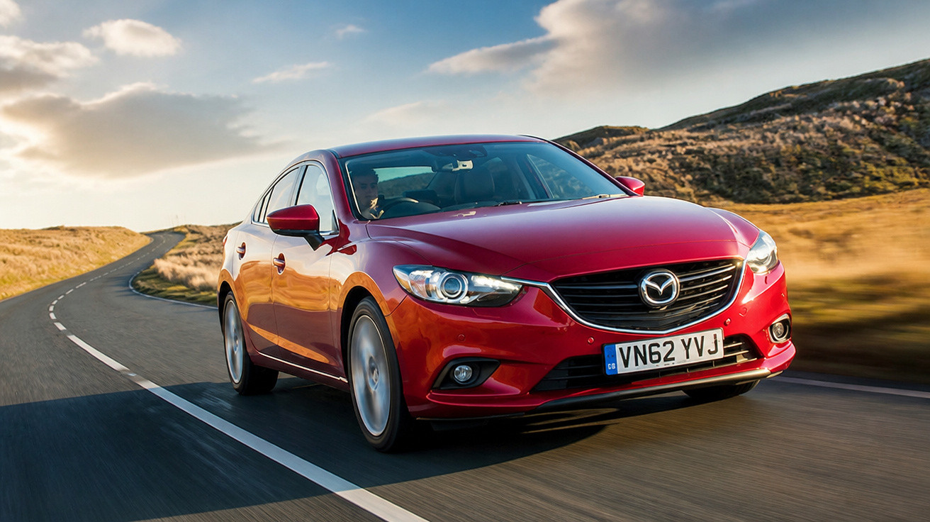 Carro sedã vermelho Mazda circulando em estrada asfaltada com paisagem de colinas ao fundo.