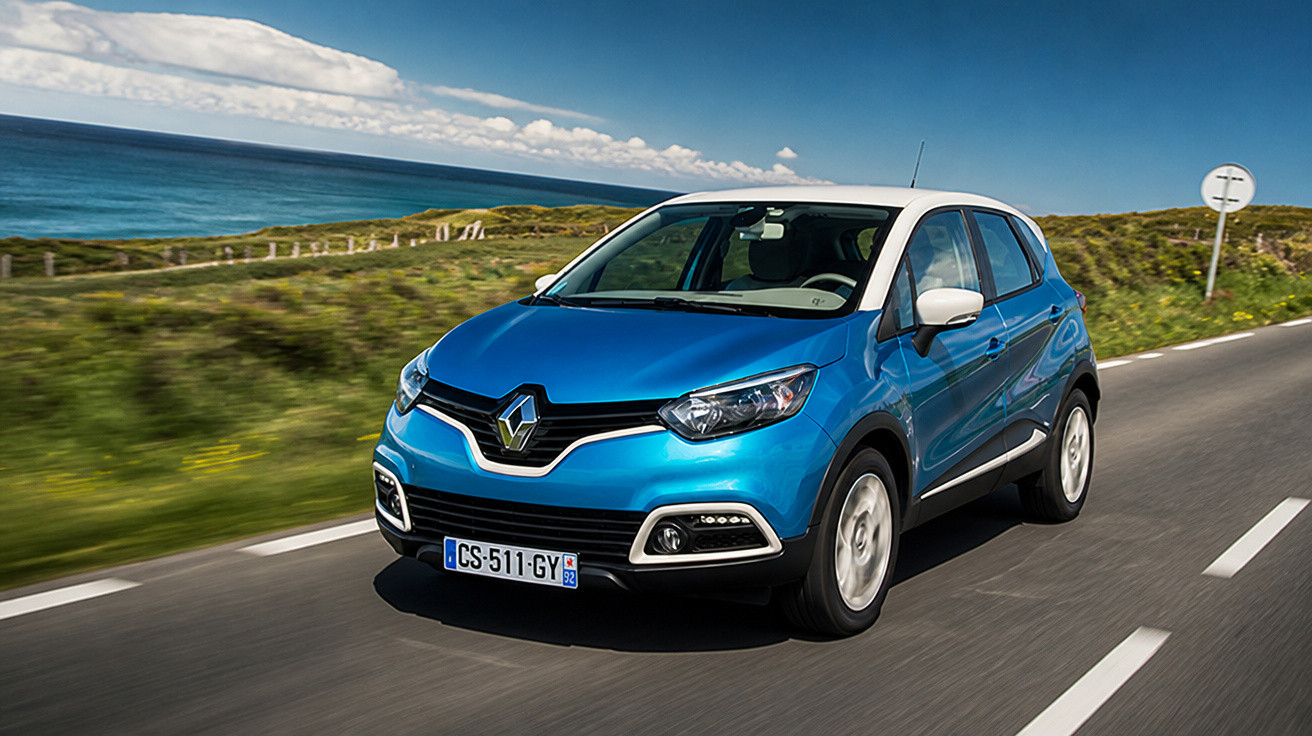 Carro Renault azul com teto branco dirigindo em estrada costeira com mar e colinas ao fundo.