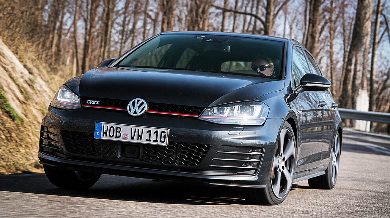 Carro Volkswagen GTI preto em estrada com árvores secas ao fundo em dia ensolarado.