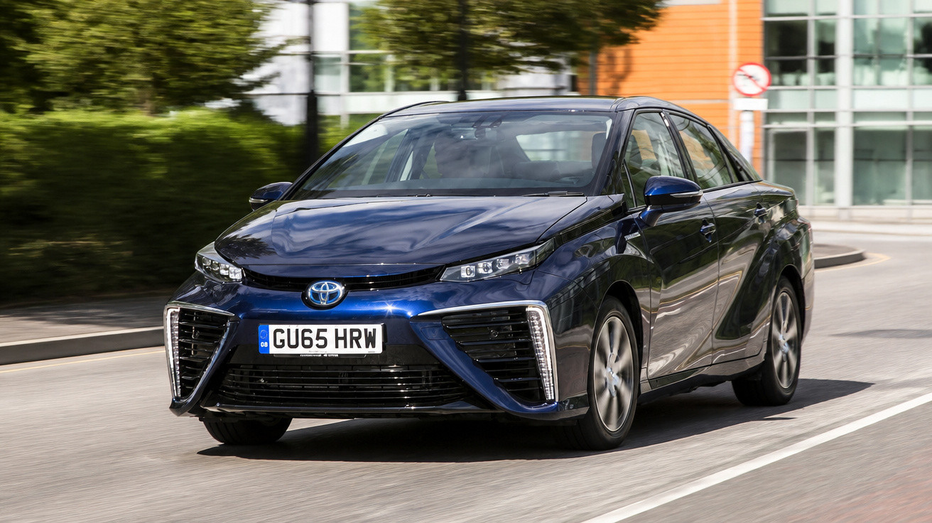 Carro Toyota azul escuro modelo sedã circulando em rua urbana com prédios e árvore ao fundo.