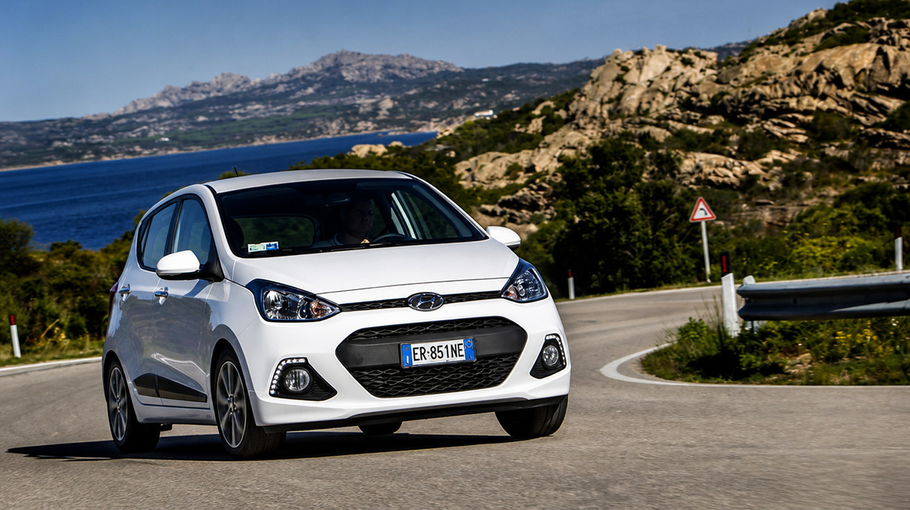 Carro branco Hyundai hatchback em curva na estrada com mar e montanhas ao fundo.