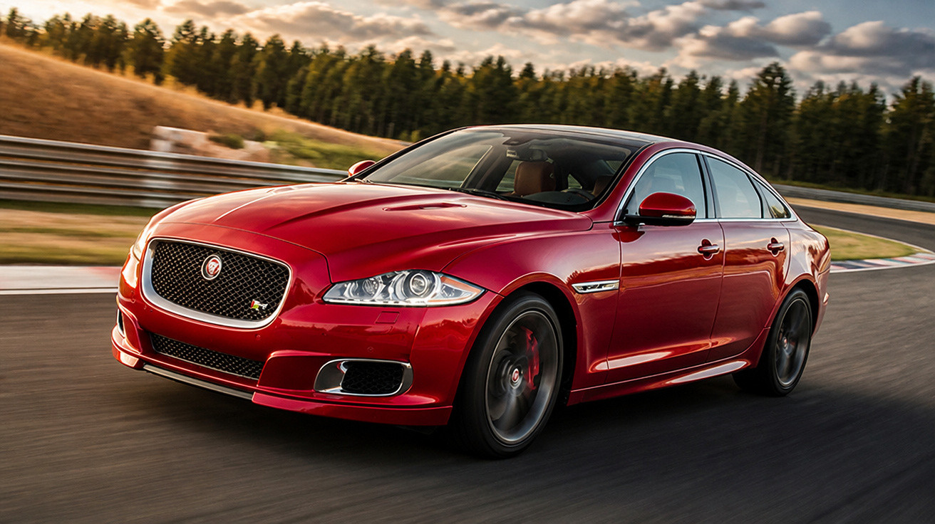 Carro esportivo vermelho Jaguar em pista de corrida com árvore e céu ao fundo.