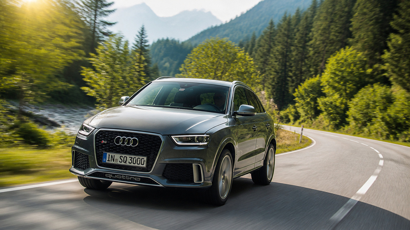 Carro Audi cinza dirigindo em estrada sinuosa rodeada por árvores verdes e montanhas ao fundo.