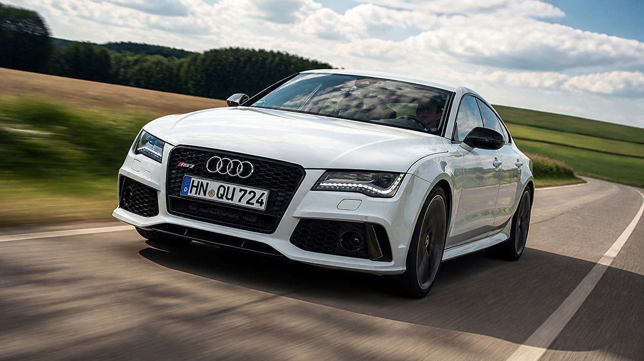 Carro branco Audi RS7 em alta velocidade em estrada sinuosa com paisagem de campos verdes ao redor.