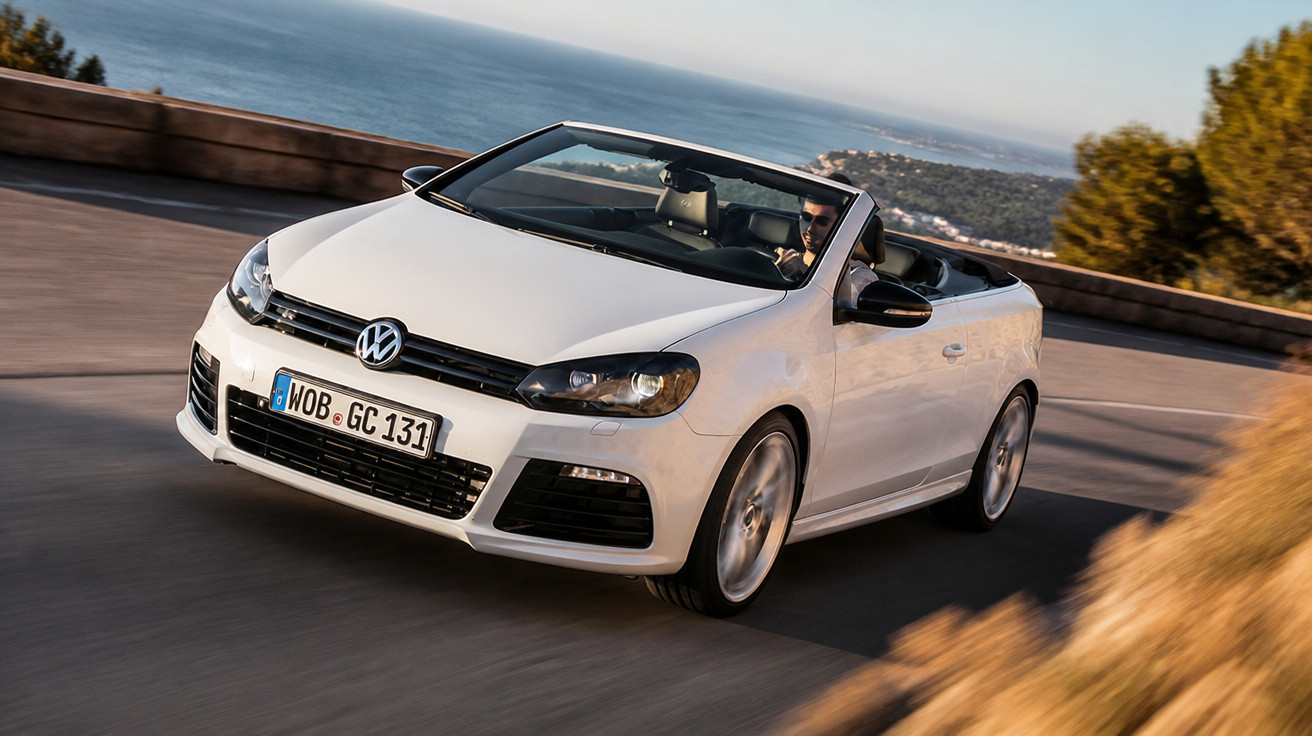 Carro conversível branco da Volkswagen dirigido em estrada costeira com vista para o mar.