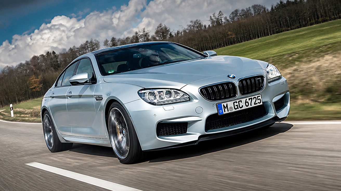 Carro esportivo BMW prata em alta velocidade em estrada com cenário de campo e árvores ao fundo.