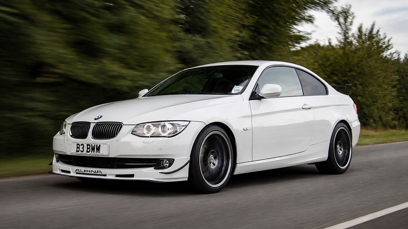 Carro BMW Alpina branco em movimento em estrada cercada por árvores verdes.